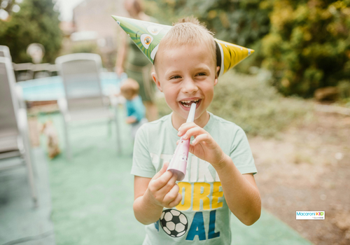young boy wearing a party hat blowing a party horn at a birthday celebration